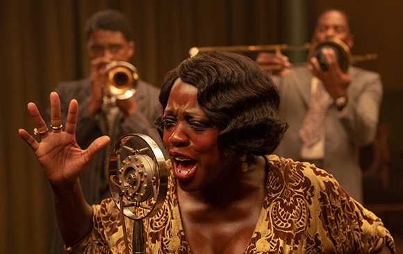 A woman sings into a vintage microphone while two men play trumpets in a dimly lit room.