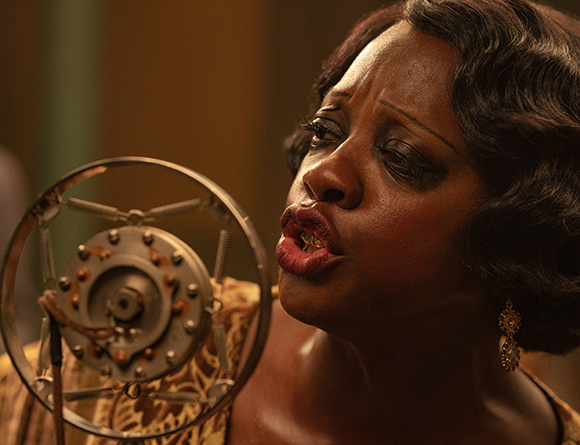A Black woman with wavy hair sings into a large, circular vintage metal microphone.