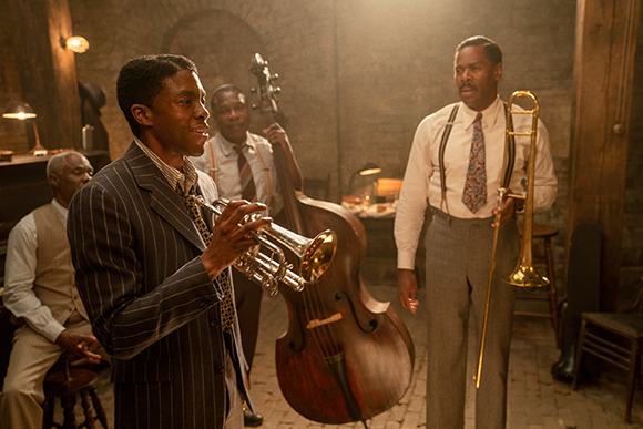 Three men in mid-century clothing pose with a trumpet, a double bass, and a trombone in a rustic room.
