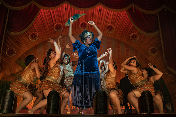 Performers in gold and blue costumes dance on a stage with red curtains and floral patterns.