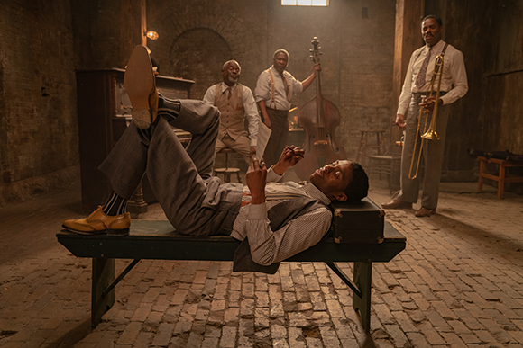 A man reclines on a bench in the foreground while three men stand with musical instruments in a brick room.