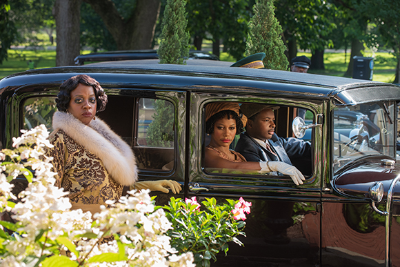 A woman in a gold patterned dress and fur collar stands next to a vintage black car in a park.