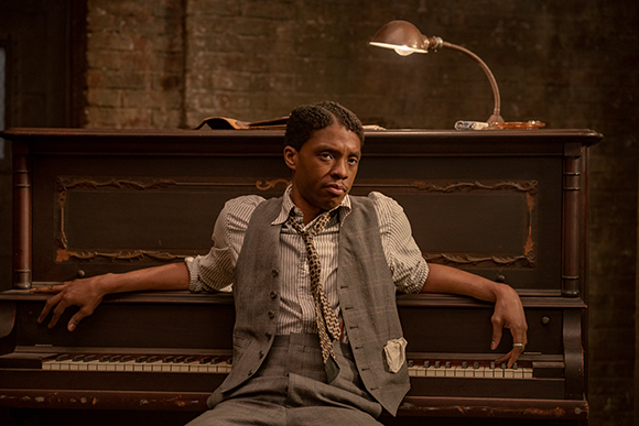 A man in a grey vest and patterned tie sits leaning against a dark wooden upright piano.