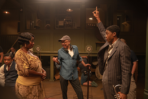 A woman in a lace dress, a man in a blue shirt and cap, and a man in a pinstripe suit pose in a studio.