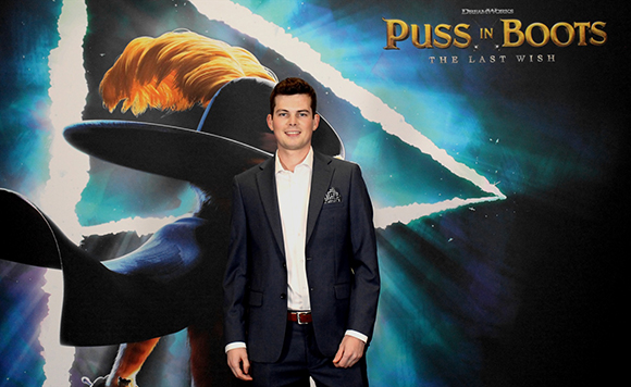 A man in a dark suit and white shirt stands before a large Puss in Boots: The Last Wish movie poster.