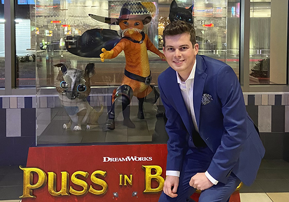 A man in a blue suit smiles next to a glass display featuring Puss in Boots and other animated characters.