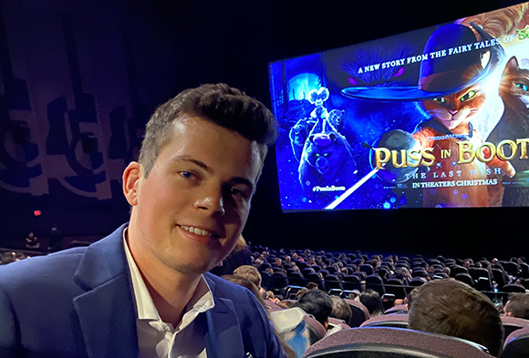 A smiling young man in a blue blazer posing in a movie theater before a Puss in Boots film.