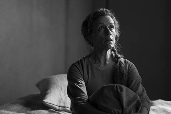 Black and white portrait of a woman with a long braid sitting on a bed and looking up.