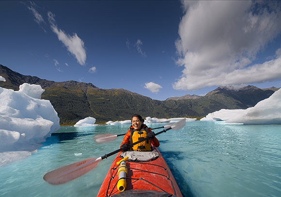 A person in an orange life vest paddles a red kayak through bright blue water surrounded by icebergs.