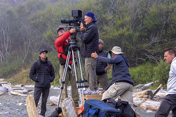 A film crew sets up a large professional camera on a tripod in a wooded, coastal area.
