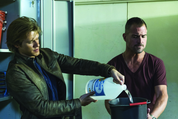 A man in a brown leather jacket pours liquid from a white jug into a black bucket held by another man.