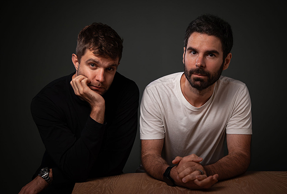 Two men sit at a wooden table against a dark background, one in black and one in white.