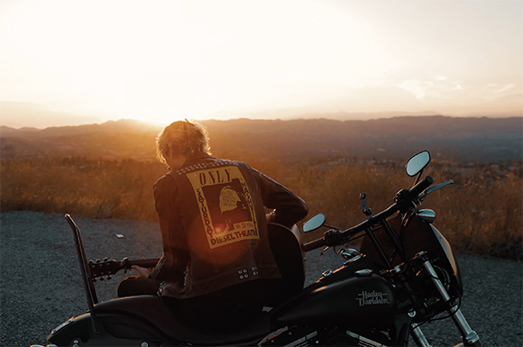 A person in a leather jacket sits on a Harley-Davidson motorcycle facing a sunset over hills.