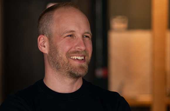 A smiling man with short blonde hair and a light beard looking off-camera.