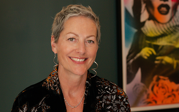 A smiling woman with short grey hair and silver hoop earrings wearing a patterned black blouse.