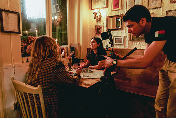 A camera operator films two women seated at a table inside a dimly lit restaurant.