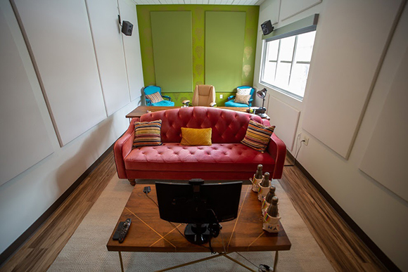 A red tufted sofa sits in a room with green walls, blue armchairs, and a wooden coffee table with a monitor.
