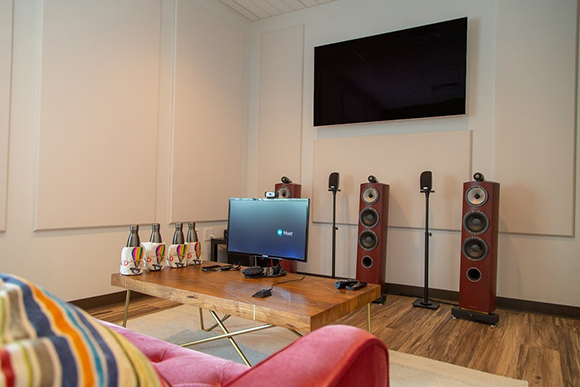 A large TV mounted on a white wall above four tall, dark wood floor-standing speakers and a computer monitor.