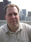 A man with short brown hair wearing a light beige button-down shirt against a city skyline.