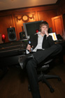 A man in a suit sits in a chair in a recording studio, holding a bottle and resting his head on his hand.