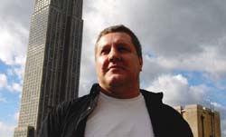 Low angle shot of a man in a white shirt and black jacket with a tall skyscraper behind him under a cloudy sky.