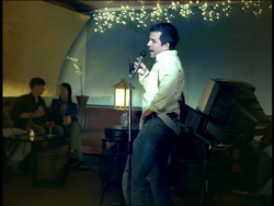 A man in a light shirt and jeans sings into a microphone on a dimly lit stage.