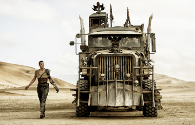 A woman in tactical gear walks across a desert toward a large, spiked, post-apocalyptic armored truck.