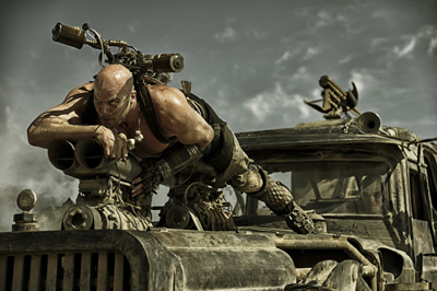 A muscular man with a shaved head crouches on a large, weathered vehicle under a cloudy sky.