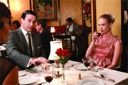 People in formal attire sitting around a white tablecloth at a restaurant table with wine and red roses.