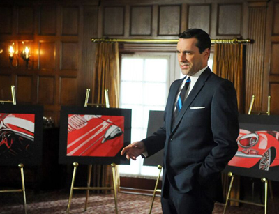 A man in a dark suit and blue tie stands in a wood-paneled room next to easels displaying red car art.