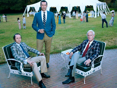 Three men in suits pose in a garden with a large white tent and party guests in the background.