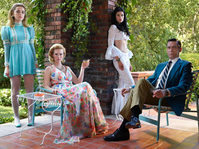 Four people in vintage attire pose in a garden near a brick pillar and patio furniture.