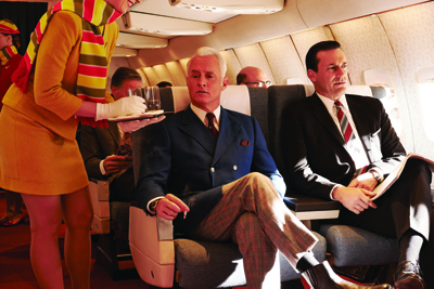 A flight attendant in a yellow uniform serves a drink to a man in a blue suit on a vintage airplane.