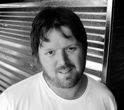 Black and white portrait of a man with dark hair and a beard wearing a white t-shirt.