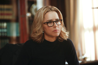 A blonde woman with black-rimmed glasses and a black turtleneck looks thoughtfully to the side.
