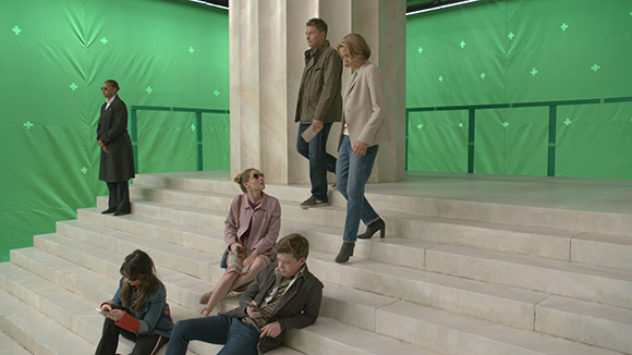 Actors standing and sitting on white marble steps in front of a large green screen.