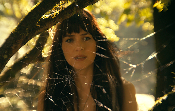 A woman with dark hair and bangs looks through a large, dew-covered spiderweb in a sunlit forest.