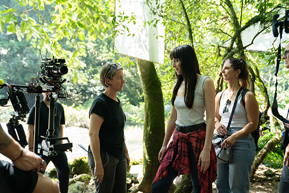 A film crew stands in a forest near a large professional camera on a rig.