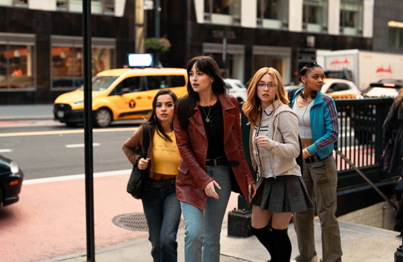 Four young women walk along a city sidewalk past a yellow taxi and urban buildings.