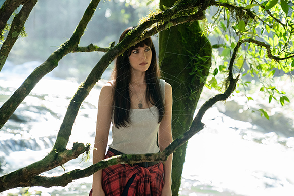 A woman with long dark hair and bangs looks off-camera behind moss-covered tree branches in a bright forest.