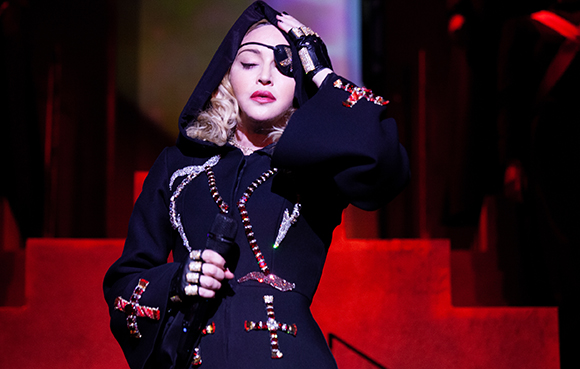 A person in a black hooded garment with silver and red cross decorations performs on stage.
