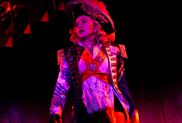 Performer in a feathered bicorne hat and decorated military jacket under dramatic red lighting.