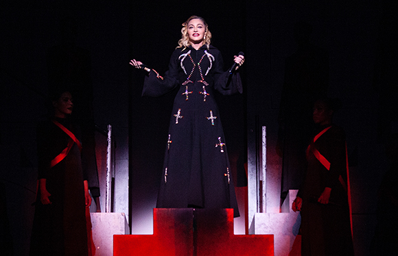 A woman in a long black robe with white embroidery stands on a stage between two other performers.