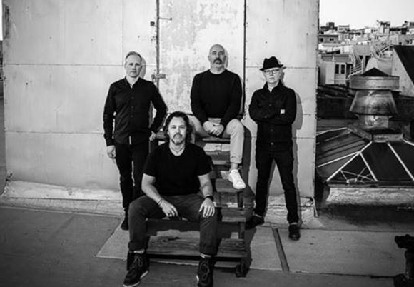 Four men in dark clothing pose on a rooftop against a light-colored wall.