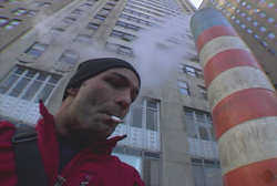 A man in a black beanie and red jacket smokes a cigarette outdoors near a tall building.