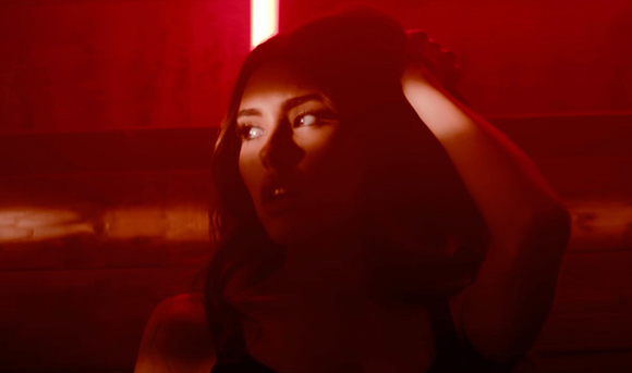 A woman with long dark hair looks to the side under intense red lighting, resting her hand on her head.