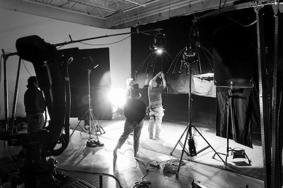 Black and white photo of a photography studio with various lights, stands, and crew members working.