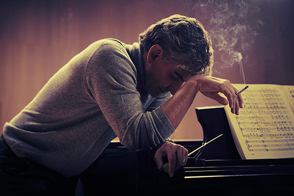 A man in a grey sweater leans over a piano, resting his head on his arm while holding a lit cigarette.