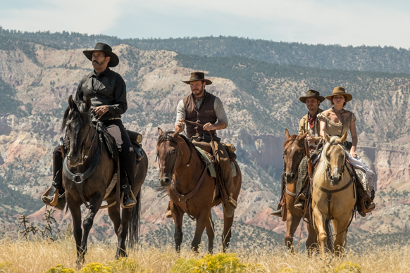 Four people in western attire riding horses across a dry, grassy field with mountains in the background.
