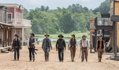 Seven men in cowboy hats and period clothing walk down a dusty street in a Western-style town.